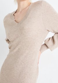 Robe pull côtelée beige avec un col en V, des manches longues, une texture douce et une silhouette ajustée, présentant des variations de couleur subtiles.