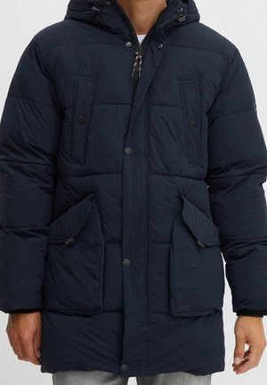 Homme portant une veste d'hiver matelassée bleu marine avec fermeture éclair devant, quatre poches à rabat et capuche, debout sur un fond uni.