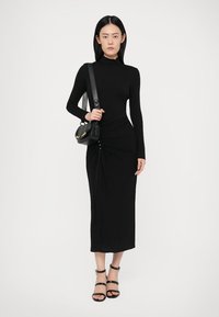 Nainen mustassa pitkähihaisessa kerrostetussa midi-mekossa, mustassa olkalaukussa ja mustissa remmihiekoissa seisoo valkoista taustaa vasten.