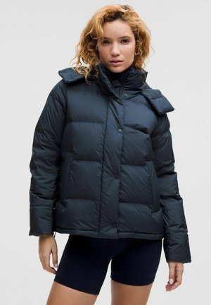 Jonge vrouw met krullend haar, gekleed in een zwarte pufferjack en zwarte strakke shorts, tegen een effen lichte achtergrond.