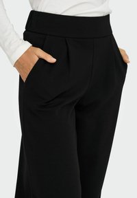 Sorte skræddersyede bukser med elastisk talje, sidelommer og pressefolder, lavet af et glat, strækbart stof. Enkel, moderne design.