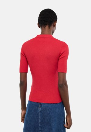 Personne portant un haut en maille rouge à manches courtes ajusté et une jupe en denim bleue, vue de dos sur un fond uni.