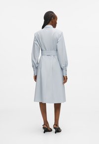 Ανοιχτό μπλε φόρεμα πουκαμίσα με ζώνη στη μέση, μακριά μανίκια, κουμπωτές μανσέτες και μήκος μίντι. Λείο ύφασμα και κλασικός σχεδιασμός γιακά.