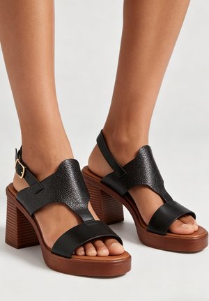 Pieds portant des sandales en cuir noir à bout ouvert, avec talons blocs en bois et brides à la cheville, sur un fond clair uni.