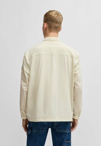 Helle beige leichte Jacke mit einem Kragen, elastischen Bündchen und einer glatten Textur. Verfügt über eine Rückenpasse und ein minimalistisches Design.