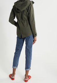 Olivgrön hooded jacka med insydd midja, uppvikta ärmar, matchad med ljusblå cropped jeans och röda flat-sandaler.