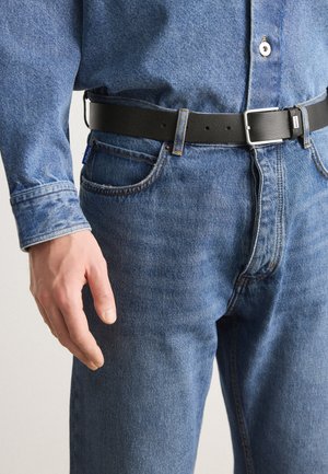 Vaqueros de denim azul claro con un corte clásico, que incluyen un cinturón de cuero negro, costuras visibles y un bolsillo trasero.