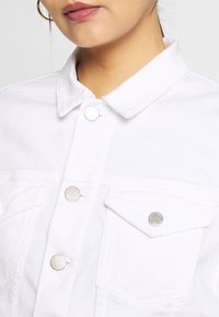 Vitt denimjacka med krage, silverfärgade tryckknappar och två bröstfickor med lock. Slät yta och strukturerad design.