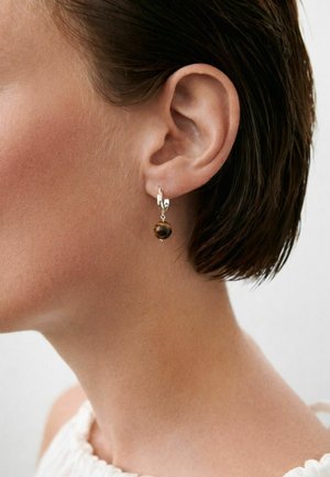 Prim-plan al urechii stângi a unei persoane cu păr scurt și închis la culoare, purtând o cercei argintiu în formă de cerc cu un pandantiv rotund întunecat.