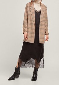 Béžový kostkovaný blazer přes černé saténové šaty s krajkovým lemováním. Šaty mají šikmý lem a jsou nošeny s černými kotníkovými botami.