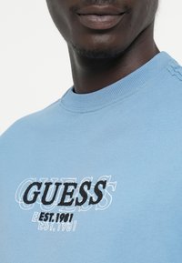 Svetlomodrá mikina s okrúhlym výstrihom, na ktorej sa nachádza slovo "GUESS" v tučných čiernych vyšívaných písmenách s vybledlým bielym okrajom.