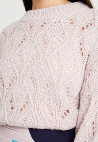Maglione lavorato a maglia rosa chiaro con un motivo a diamante, dettagli in openwork e scollo a coste. Texture morbida e design a vestibilità ampia.