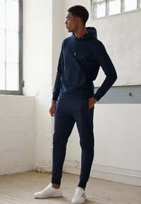 Felpa blu navy con tasca a marsupio e cordoncino regolabile, abbinata a joggers affusolati coordinati. Indossata con scarpe da ginnastica bianche.