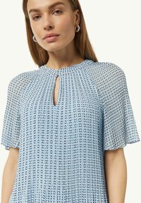 Lichtblauwe blouse van doorschijnend materiaal, met een keyhole-halslijn, korte fladdermouwen en een geometrisch patroon in donkerdere blauwtinten.