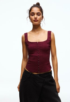 Woman with hair in a bun wearing a sleeveless burgundy top with front hook closures and oversized black pants against a plain white background.