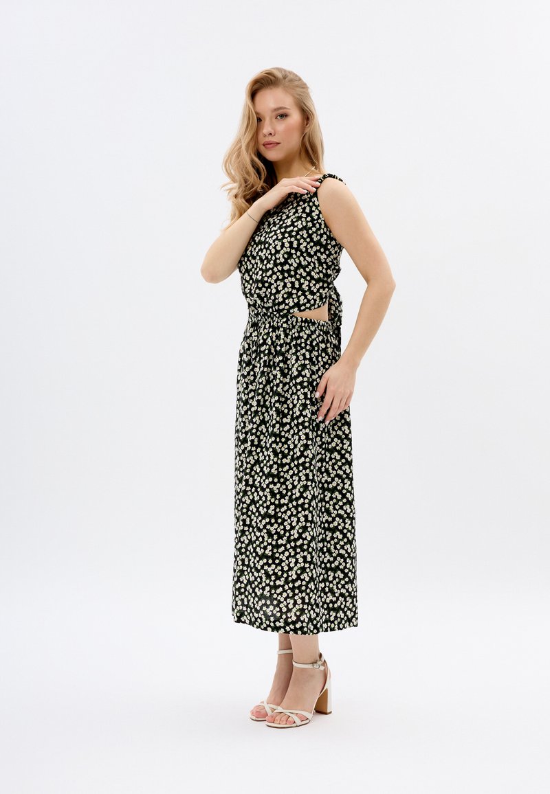 Schwarzes Blumenkleid im Maxistil mit weißem Gänseblümchenmuster, Cut-Out-Taille und ärmellosem Design. Kombiniert mit hellen, verschnürten Absätzen.