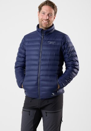 Glunderende man in een blauwe gewatteerde Swedemount-jack met zijn handen in de zakken en zwarte outdoor broek met rits tegen een eenvoudige achtergrond.