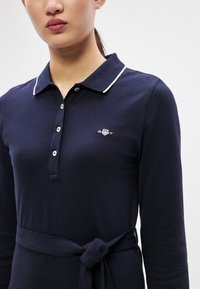 Tumši zila polo kleita ar garām piedurknēm un baltu apkakli, ar pogājamu plāksteri un sasienamu jostasvietu. Uz krūtīm izšūts Gant logo.