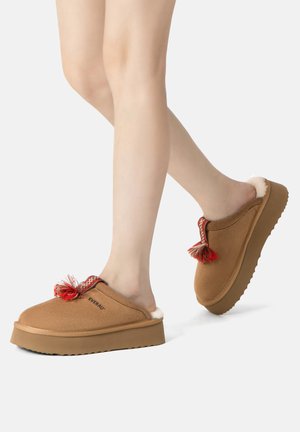 Braune Wildleder-Clogs mit einem flauschigen weißen Futter, versehen mit einer dekorativen roten und weißen Quaste sowie einer strukturierten Gummisohle für besseren Halt.