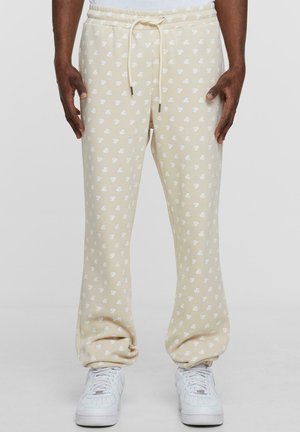 Beige sweatpants with an all-over white logo pattern, relaxed fit, elastic waistband with drawstrings, and side pockets.