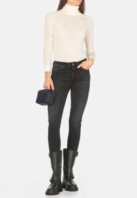 Maglione bianco a collo alto, jeans skinny scuri e stivaletti neri alla caviglia. Una piccola borsa blu navy è tenuta in una mano.