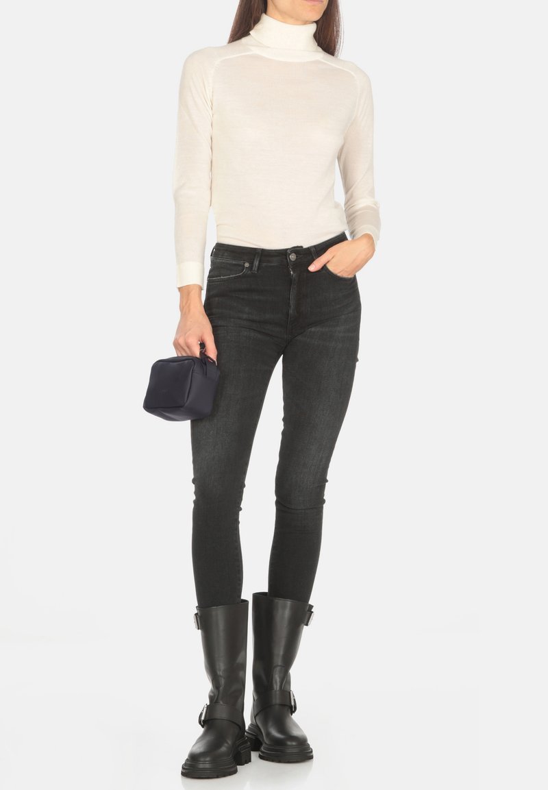 Maglione bianco a collo alto, jeans skinny scuri e stivaletti neri alla caviglia. Una piccola borsa blu navy è tenuta in una mano.