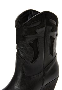Bota de cowboy de cuero negro con costuras decorativas y un tacón de cuña de altura media, vista desde un ángulo lateral en primer plano.