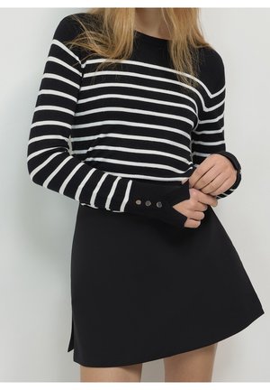 Femme portant un haut à manches longues rayé noir et blanc avec des trous pour le pouce et des boutons, associé à une jupe noire.