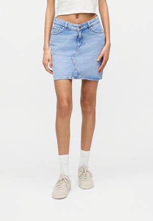 Femme portant une minijupe en jean bleu clair, un haut court blanc, des chaussettes blanches et des baskets beige, debout devant un fond uni.