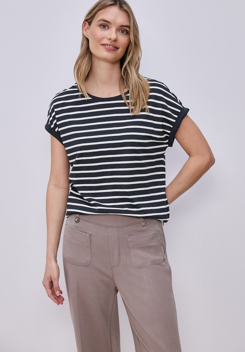 Mujer con camiseta de manga corta a rayas blancas y negras y pantalones topo, de pie con una mano en la cadera sobre un fondo liso.