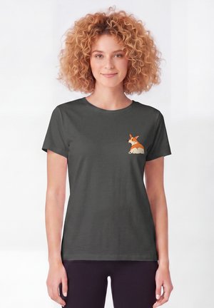 Femme aux cheveux bouclés portant un t-shirt gris foncé avec un petit motif graphique d'un chien corgi orange et blanc sur la poitrine gauche.