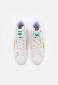 Bílé vysoké tenisky Puma s oranžovými bočními pruhy a zeleným logem Puma na jazyku, zobrazené zeshora na bílém pozadí.