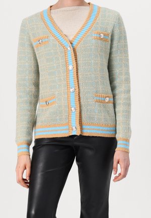 Cardigan de punto verde claro con rayas azules y beige, botones plateados, cuatro bolsillos, llevado sobre una blusa beige y pantalones de cuero negro.