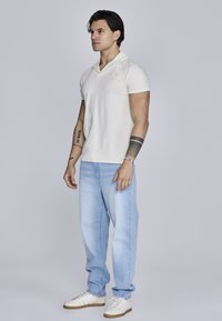 Creme polo skjorte, lyseblå jeans og hvide sneakers; har korte ærmer og en afslappet pasform med minimal branding.