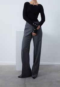 Haut noir à manches longues avec des boutons dorés au niveau de l'encolure et des poignets, associé à un pantalon gris taille haute à jambes larges, et des chaussures noires pointues.