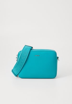 Petit sac bandoulière rectangulaire turquoise avec une large sangle embossée du logo Calvin Klein sur un fond blanc uni.