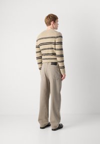 Beige villatakki mustilla raidoilla, tekstuurikuvioitu neule. Vaaleanharmaat leveälahkeiset housut takataskuilla. Malli selkä menosuuntaan, seisoo neutraalilla taustalla.