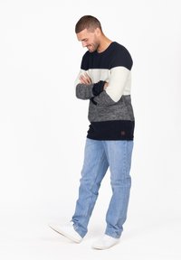 Maglione a righe in navy, crema e grigio a maglia. Presenta maniche lunghe e scollatura a girocollo. Abbinato a jeans azzurri e sneakers bianche.