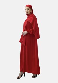 Rød, langærmet kjole og matchende hijab med samlet ærmer; fremhævet med guldtrim; båret med sorte højhælede sko.