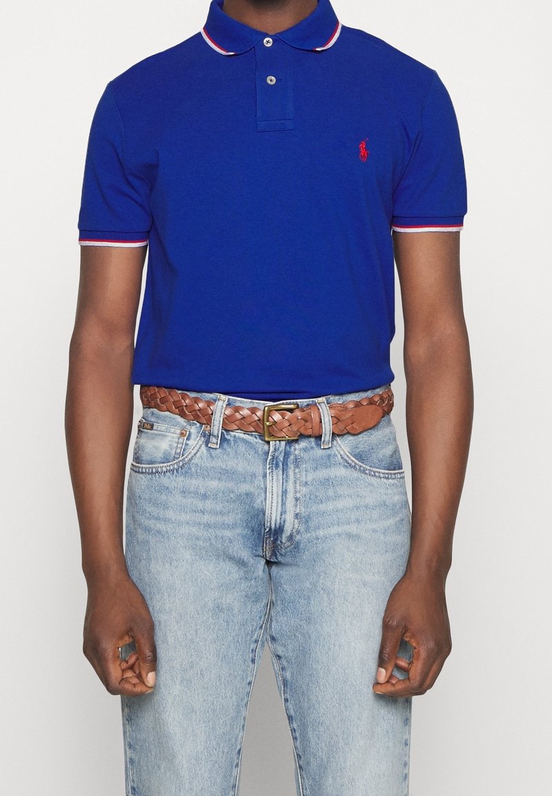 Homme portant un polo bleu avec des bordures rouges et blanches, un jean bleu clair et une ceinture en cuir tressé marron avec une boucle en laiton.