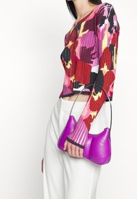 Fialová kožená kabelka s jedinečným zakřiveným tvarem a tenkým černým ramenním popruhem, kombinovaná s pestrobarevným žebrovaným dlouhým rukávem.