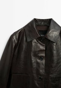 Veste en cuir noire avec grand col et trois boutons sur le devant, montrant une texture lisse et deux poches avant.