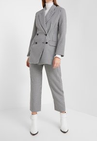 Donna che indossa un blazer doppiopetto a scacchi bianco e nero e pantaloni cropped coordinati, con stivaletti bianchi su uno sfondo bianco.