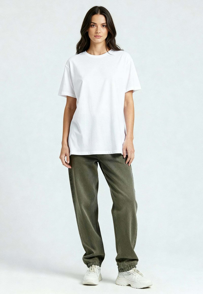 Jeune femme debout, face à l'avant, portant un t-shirt blanc uni, un pantalon vert olive et des baskets blanches, sur un fond clair.