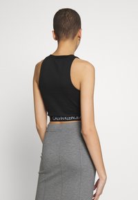 Person som bär en svart kortärmad topp med "Calvin Klein Jeans"-band och en högmidjad grå blyertskjol, vänd bort mot en vit bakgrund.