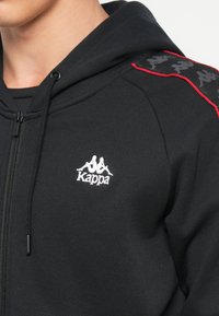 Sudadera con cremallera negra hecha de un tejido suave, con un logo blanco de Kappa y detalles rojos en los hombros con una franja negra con patrón.