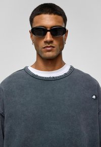 Donkergrijze sweatshirt met een versleten halslijn en een klein gaatje-detail, gedragen over een witte shirt. Zwarte ovale zonnebril maakt de look compleet.