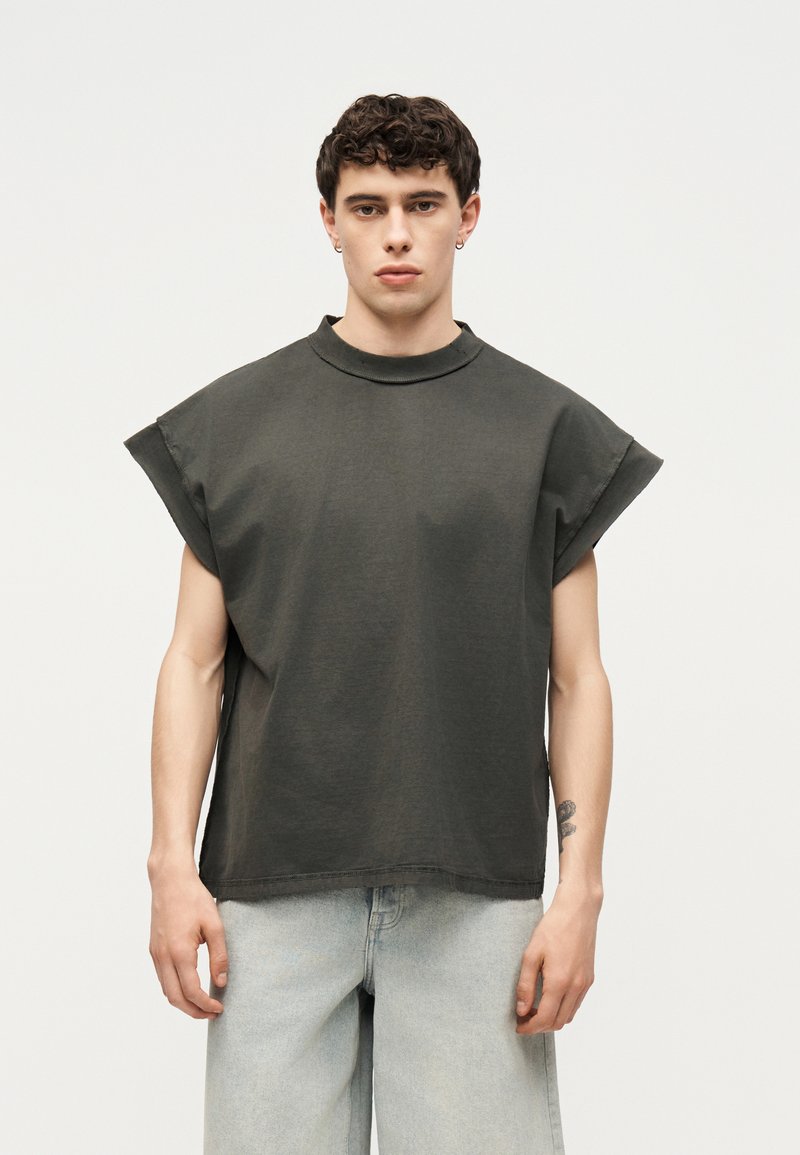 Νέος άνδρας με κοντά κατσαρά μαλλιά, φοράει σκούρο αμάνικο μπλουζάκι και ανοιχτό μπλε τζιν, στέκεται μπροστά σε απλό φόντο.