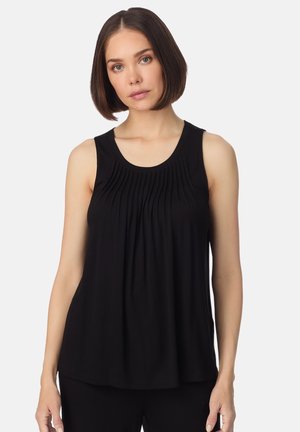 Frau mit kurzen braunen Haaren, die ein ärmelloses schwarzes Oberteil mit vertikalen Falten auf der Brust trägt, steht vor einem weißen Hintergrund.