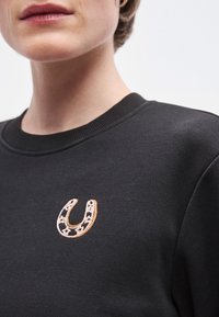 Μαύρο φούτερ με στρογγυλή λαιμόκοψη και κεντημένο λογότυπο πετάλου σε πορτοκαλί και λευκό στο μπροστινό μέρος. Μαλακή υφή, casual σχεδιασμός.